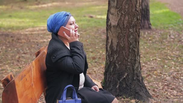 Eine Jüdin mit traditioneller Kopfbedeckung im Profil auf einer Bank in einem herbstlichen Park telefoniert — Stockvideo