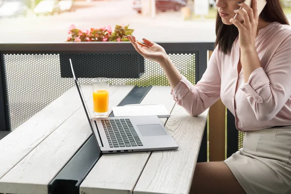 Jovem mulher de negócios falando ao telefone e gesticulando segurando um lápis, laptop e suco na mesa . — Fotografia de Stock