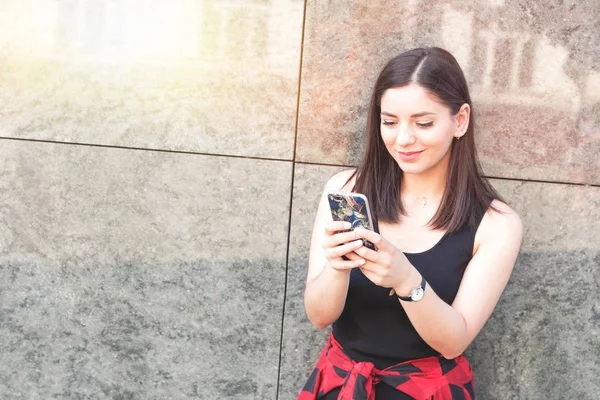 Chica feliz mensajes de texto en un teléfono inteligente apoyado en la pared de mármol en la ciudad . — Foto de Stock