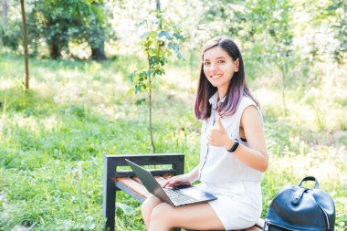 Parkta dizüstü bilgisayarda uzaktan öğrenen neşeli genç öğrenci sosyal mesafeyi koruyor ve yalnız zaman geçirmenin tadını çıkarıyor.