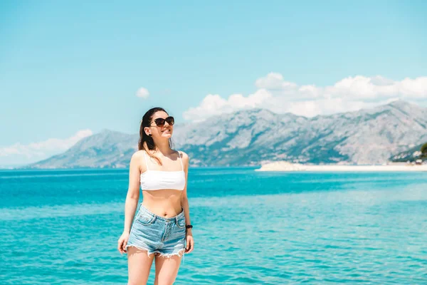 freedom of travel. beautiful young woman alone along blue sea with mountains on background looking in the distance sunglasses on smiling and enjoying travel.