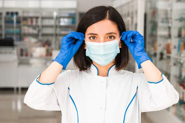 Retrato próximo farmacêutico feminino na farmácia colocando no rosto uma máscara protetora chegando ao trabalho para ajudar as pessoas — Fotografia de Stock