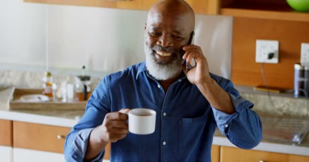 を自宅の台所で携帯電話で話しながらブラック コーヒーを持つ年配の男性 — ストック動画