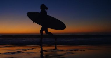 Kadın sörfçü surfboard ile 4 k gün batımında sahilde yürüyüş
