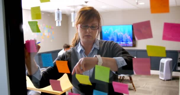 Female Executive Using Smartwatch While Writing Sticky Note Glass Wall — Stock Video