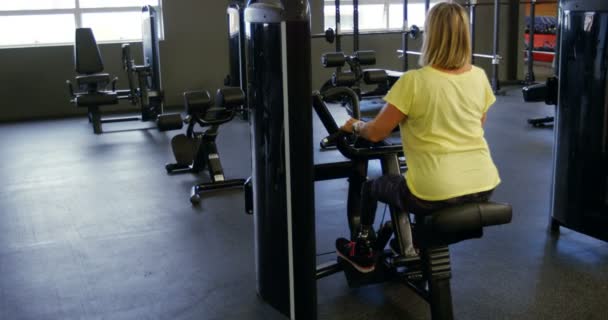在健身室4K 的机器上做背部运动的高级妇女的后方视图 — 图库视频影像
