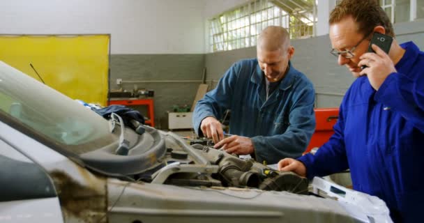 Mecánico Examinando Coche Mientras Colega Habla Teléfono Móvil Garaje Reparación — Vídeos de Stock