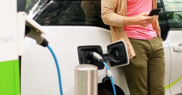 Mittelteil Des Menschen Mit Handy Beim Laden Von Elektroauto Ladestation — Stockvideo