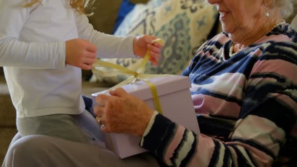 側ビューの素敵な孫娘は 自宅のリビング ルームで彼女の祖母に贈り物を与えます 彼らは贈り物を Unbox に黄色いリボンを元に戻す — ストック動画