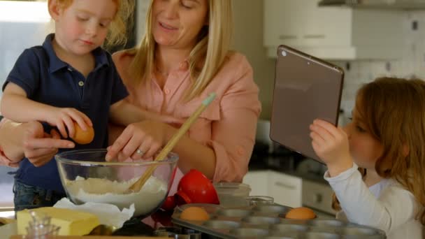 母親と自宅のキッチンで食事を準備する子どもたちの正面から見た図 母は彼女の娘にケーキを焼くために勉強します 一人の小さな女の子が速報卵とタブレットでレシピを探して — ストック動画