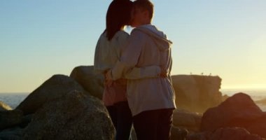 Birbirimizi kayalık sahilde Öpüşme Romantik Çift yan görünüm. 4k arka planda gökyüzü günbatımı