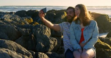 Mutlu genç çift sahilde kayaya selfie tıklayarak gülümseyerek. Tur 4 k oturan kadın adamın