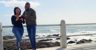 Mutlu çift cep telefonuyla korkuluk yakınındaki güneşli bir günde. Güzel gök ve 4k planda sakin deniz