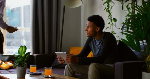 在舒适的家中 在沙发上使用数字平板电脑的混血年轻人的正面景观 混血年轻女子带着盘子进入 坐下来与男子在数字平板电脑4K 上进行讨论 — 图库视频影像