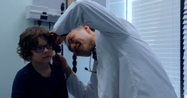 クリニックでツールで白人少年患者耳を調べる若いアジア男性医師の側面図です 男性医師患者 との相互作用 — ストック動画