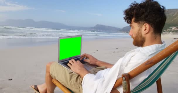 若い白人男ラウンジチェアに座っていると ビーチでノート パソコンを使用しての側面図です 晴れた日に のビーチでリラックスした男 — ストック動画