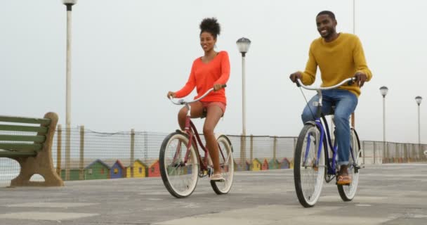 晴れた日のビーチの遊歩道に黒の若いカップル乗馬の自転車のフロント ビュー カップルの笑顔と楽しいビーチ — ストック動画