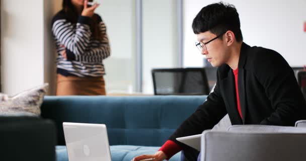 亚洲商人在笔记本电脑上工作的侧视图 并在现代办公室的记事本上写作 在后台4K 的手机上交谈的女性同事 — 图库视频影像