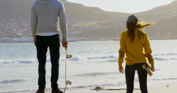 年轻的高加索滑板运动员站在海滩上看大海的后景 他们在阳光下拿着滑板 — 图库视频影像