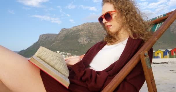 サンラウン ジャーの上に座って ビーチで本を読んでのサングラスをかけた若い白人女性の側面図です の晴れた日にビーチでリラックスできる女性 — ストック動画