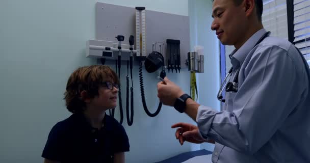 クリニックで白人少年患者へ注射器を示す若いアジア男性医師の側面図です 男性医師患者 との相互作用 — ストック動画