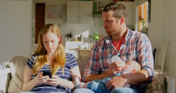 彼の赤ちゃんと母親の家で携帯電話を使用してミルクを授乳アダルト白人父半ば正面 お互い を探している親 — ストック動画