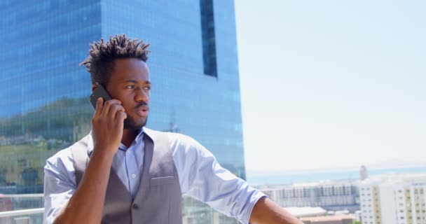 近代的なオフィスのバルコニーで携帯電話で話している若い黒人実業家のフロント ビュー 晴れた のバルコニーに立つビジネスマン — ストック動画