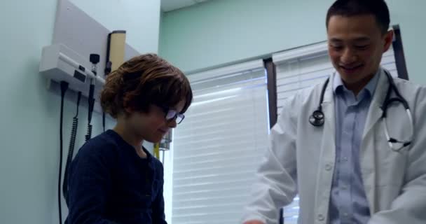 クリニックで白人少年患者の膝を調べる若いアジア男性医師のフロント ビュー 反射ハンマー を使用して男性医師 — ストック動画
