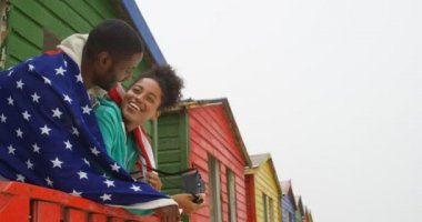 Amerikan bayrağı sarılı ve plaj kulübe, Korkuluk üzerine eğilerek genç siyah çift yan görünüm. Gülümseyen ve 4 k birbirleri ile etkileşim