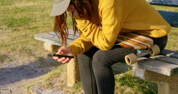 音楽を聴くと ビーチで携帯電話を使用しての若い白人女性の側面図です サンシャイン の木製ベンチのスケート ボードと座っている女性 — ストック動画