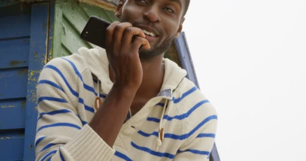 在阳光明媚的日子里 年轻黑人在海滩上用手机说话的正面景色 男人微笑着 环顾四周4K — 图库视频影像