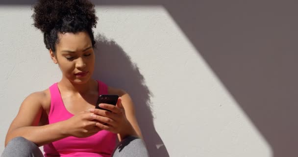 家の裏庭で壁に座っている若い混血女性の正面から見た図 彼女は 太陽の光で携帯電話にテキスト メッセージです — ストック動画