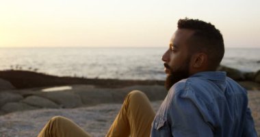 Düşünceli karışık ırk kayada kumsalda günbatımı sırasında oturan adam. karışık ırk adam denizde seyir 4 k