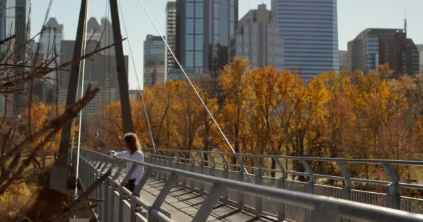 街の橋の上のコーヒーを飲んでいる若い白人男性の側面図です 高層ビルと 都市の建物の背後にある秋の木々 — ストック動画