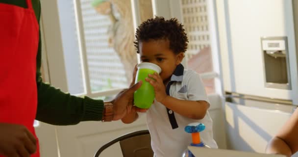 快適な家の台所で息子に水を供給する黒人の父の中間セクション の息子を見て母親 — ストック動画