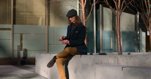 年轻的白种人在城市使用手机的侧视图 英俊的人坐在周围的墙壁4K — 图库视频影像