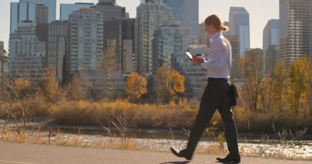 年轻的白种人在城市的道路上行走时使用手机的侧视图 阳光明媚的天气4K 的现代高层建筑 — 图库视频影像