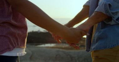 Karışık yarış çift elele duran kaya Plajı'nda orta bölümü. Romantik karışık ırk çift ayakta günbatımı 4k sırasında plajda