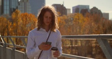 Genç Beyaz adam cep telefonuyla köprüde önden görünümü. Güneşli bir günde 4k şehir köprüsü üzerinde tek kullanımlık kahve Kupası duran ile gülümseyen adam