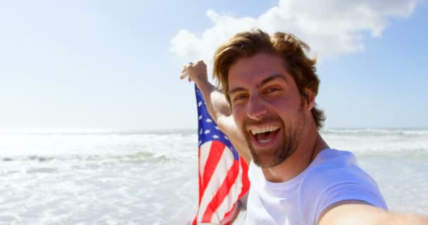 若い白人カップルのビーチにアメリカの国旗を振っている保有物の側面図です のビーチで楽しんでください — ストック動画