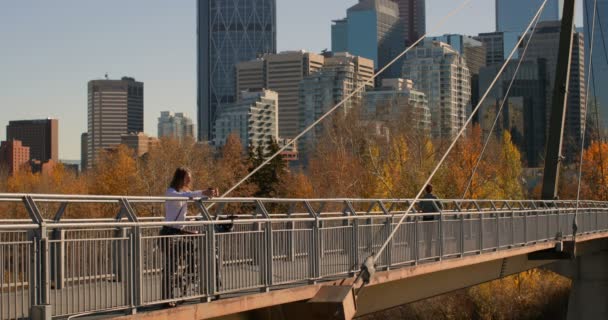 街の橋の上のコーヒーを飲んでいる若い白人男性の側面図です 高層ビルと 都市の建物の背後にある秋の木々 — ストック動画