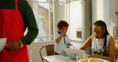 Önden Görünümü çalışma dizüstü ve mutfakta yemek masasında oynarken oğlu siyah genç anne. Ayakta ve kase 4 k tutan babası