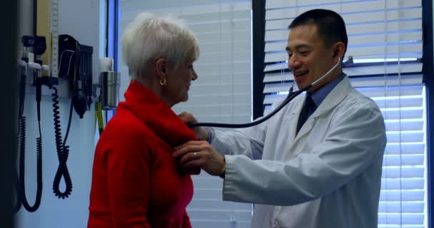 シニア クリニックで検査若いアジア男性医師の側面図です 若いアジア男性医師患者 シニアとの対話 — ストック動画