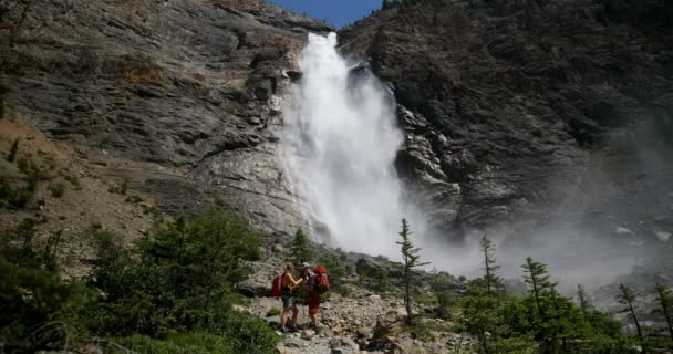 在阳光明媚的日子里 年轻的高加索徒步者夫妇的侧视图与背包站在森林里 美丽的瀑布在背景4K — 图库视频影像