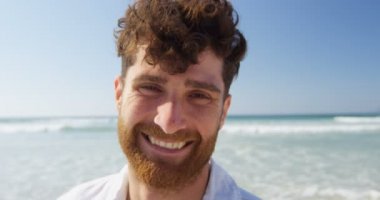 Mutlu genç adam sahilde kamera bakıyor. Mutlu adam güneşli bir günde 4 k kumsalda ayakta