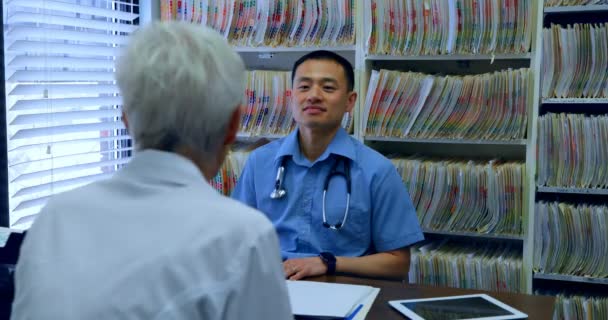 若いアジアの男性医師と先輩患者の診療所で お互い相互作用します 若いアジア男性医師と の机に座ってシニアの患者 — ストック動画