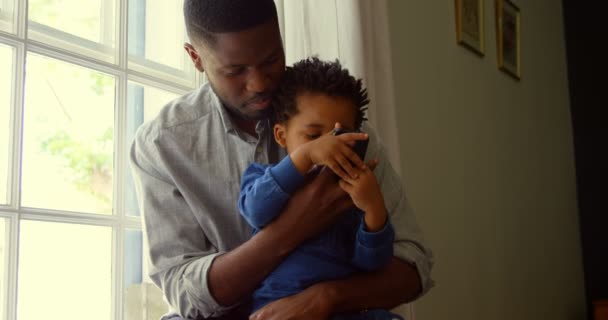黒人の父と快適な家でスマート フォンを使用して息子の正面 スマート フォン でテキスト メッセージ — ストック動画