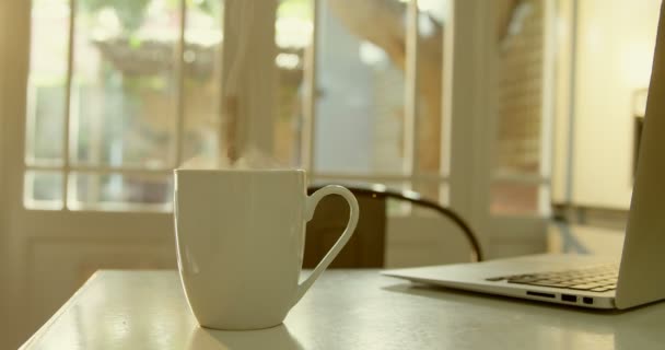 コーヒーと快適な家の台所にダイニング テーブルの上のノート パソコンのマグカップのクローズ アップ コーヒーのマグカップ から出る蒸気 — ストック動画
