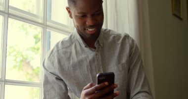 Önden Görünümü smartphone kullanarak ve pencere pervazına konforlu bir evde oturan genç siyah adam. O gülümseyen ve 4 k cep telefonunda mesaj.