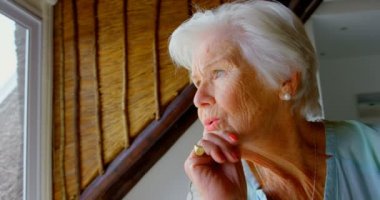 Pencere evde oturan düşünceli eski beyaz kadın kıdemli yan görünüm. O pencereden 4 k yağma